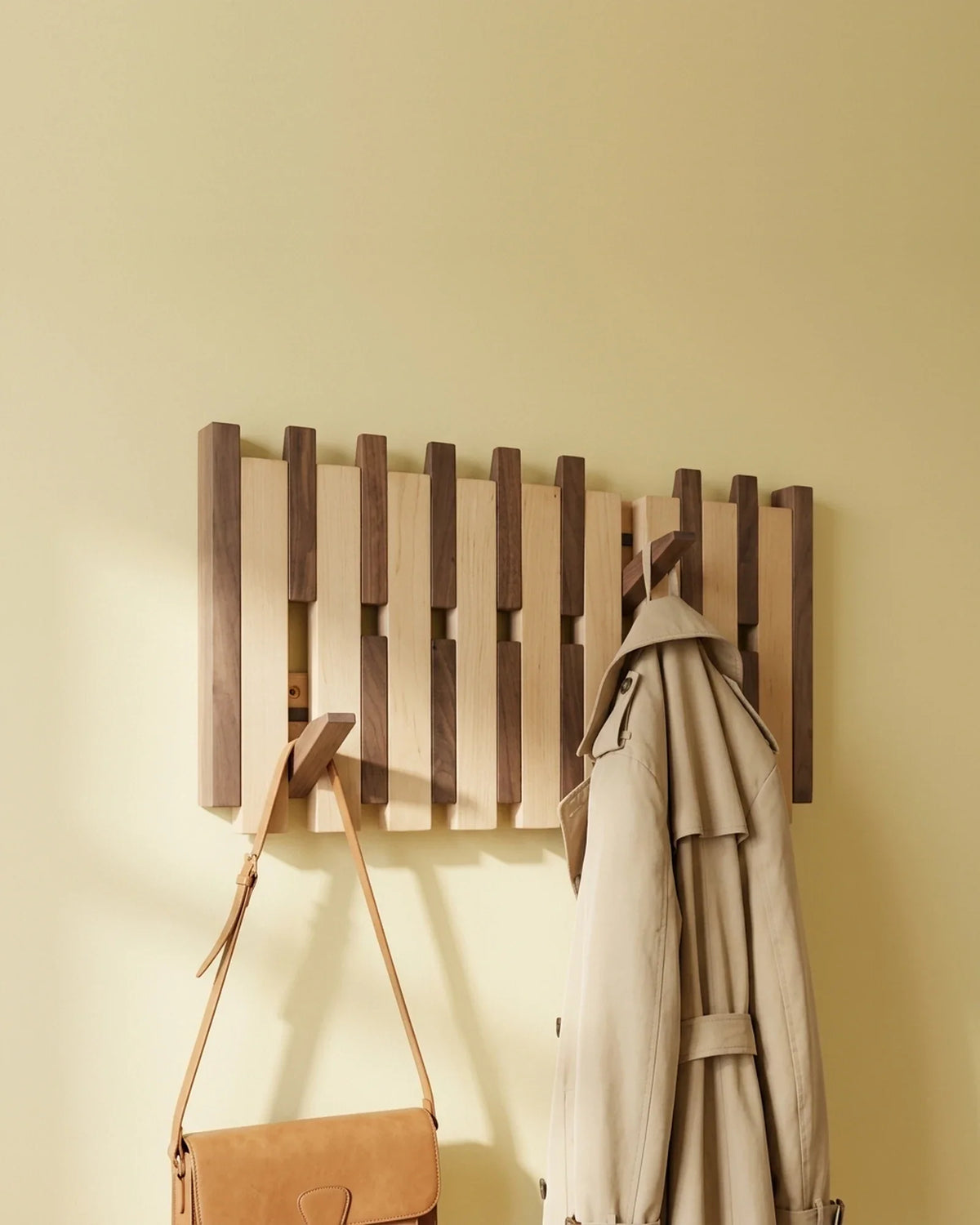 Two-tone piano key coat rack on a primrose yellow wall, holding a leather satchel and a beige trench coat