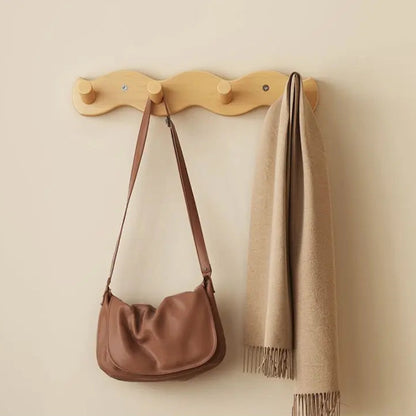 A light natural wood wall-mounted coat rack with four hooks, shown hanging a brown shoulder bag and a beige scarf against a warm beige wall. Chinese text appears along the bottom of the image.