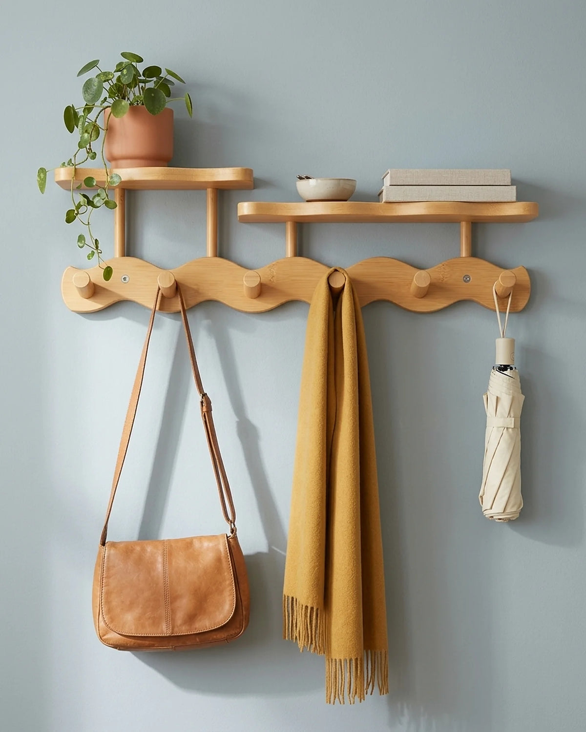 Natural bamboo wavy coat rack with two shelves holding a plant, books, a leather bag, a mustard scarf and an umbrella on a blue-grey wall
