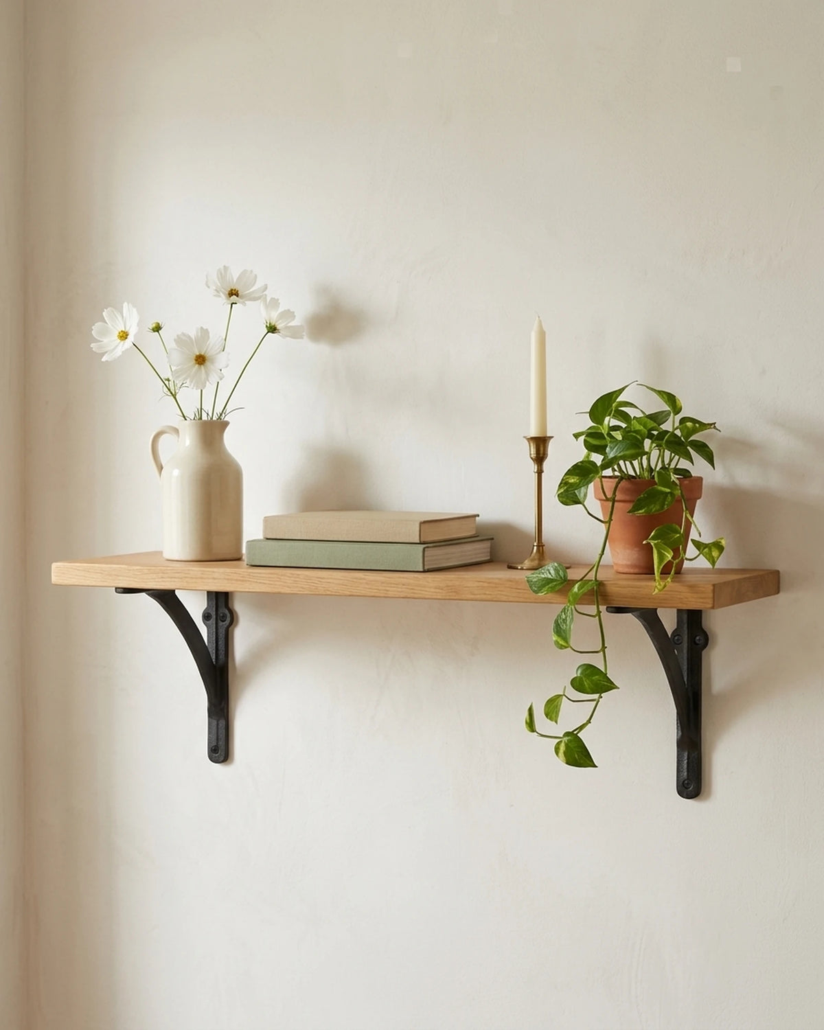 Arc black cast iron shelf brackets holding an oak shelf with books, a jug of cosmos and a brass candlestick on a warm white wall