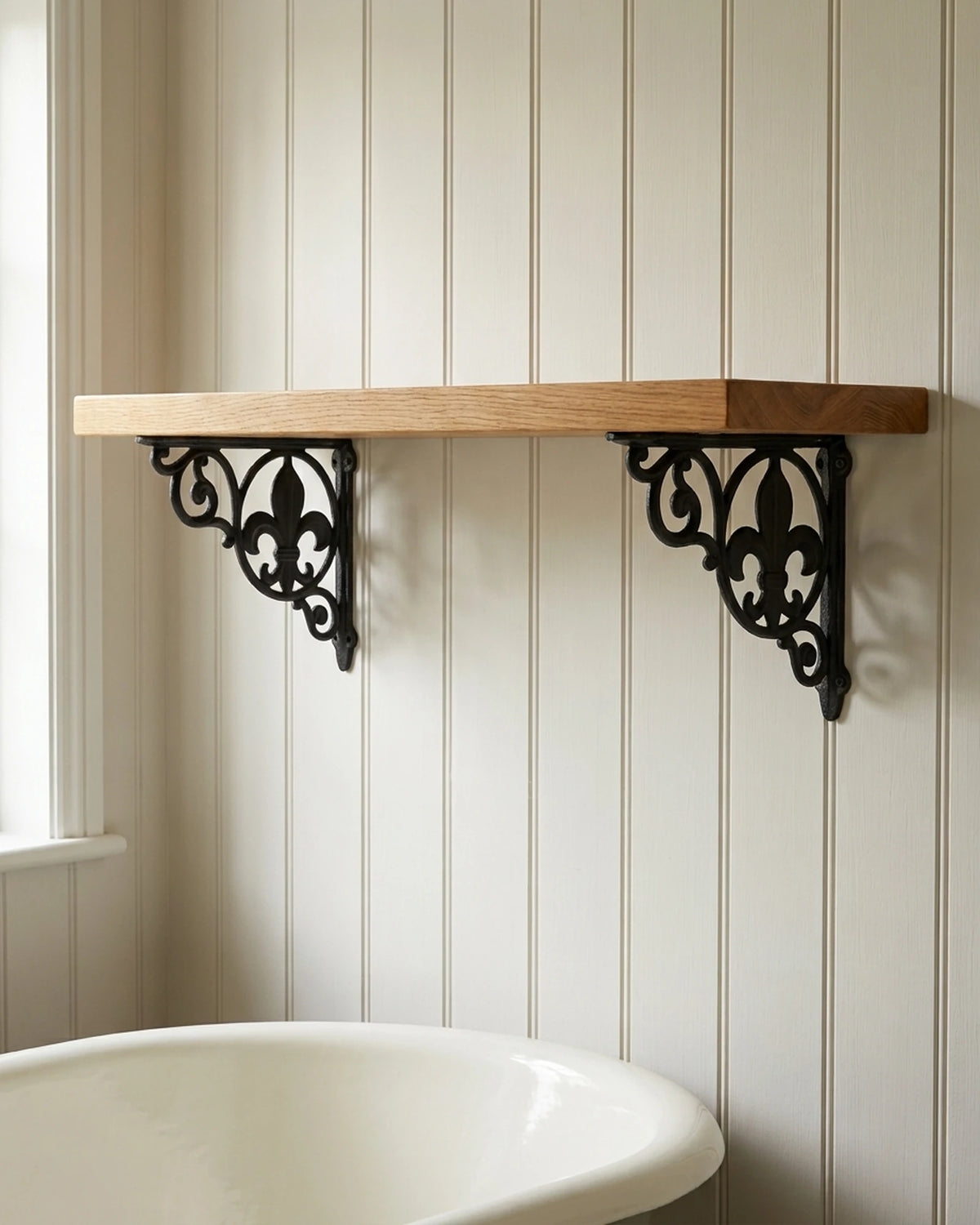 Spear Lace black cast iron shelf brackets with a fleur-de-lis holding an empty oak shelf in an off-white cottage bathroom