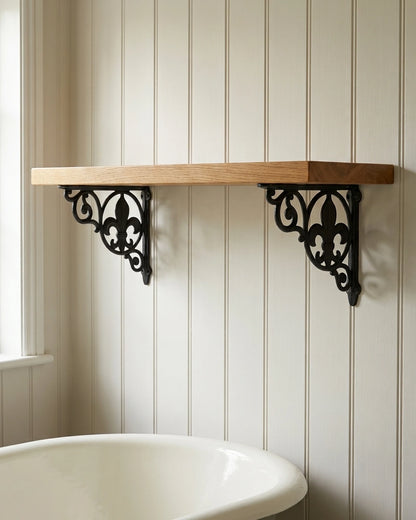 Spear Lace black cast iron shelf brackets with a fleur-de-lis holding an empty oak shelf in an off-white cottage bathroom