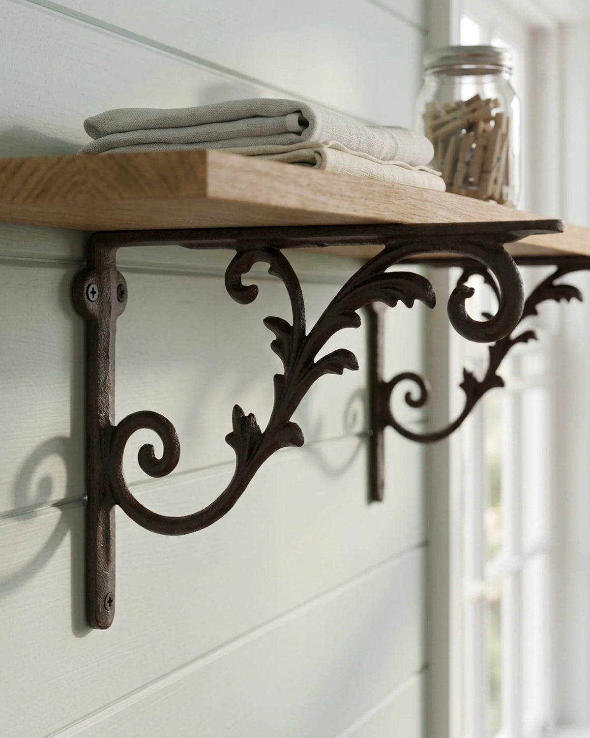 Close-up of a light scroll decorative shelf bracket holding an oak shelf on a pale celadon shiplap wall in a cottage laundry room