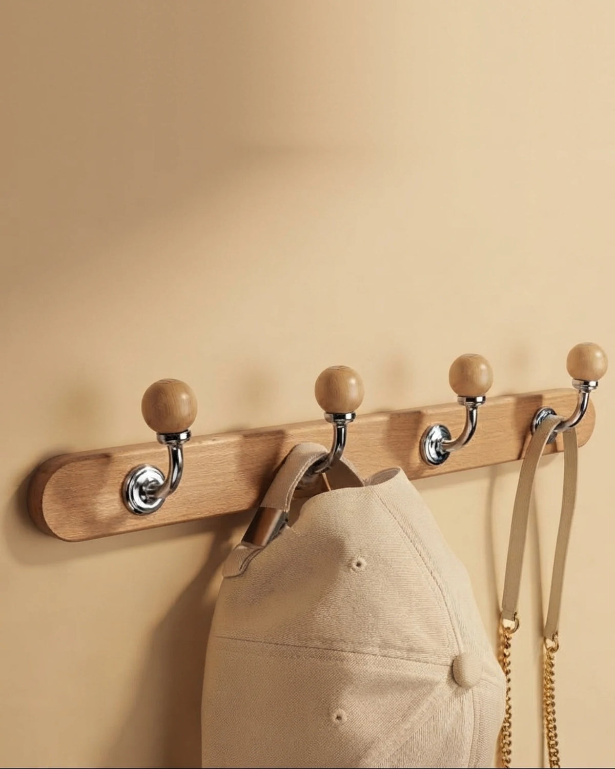 Natural wood coat rack with four chrome ball-top hooks on a warm beige wall, holding a cotton cap and a shoulder bag with a gold chain strap