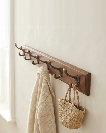 Wooden coat rack with six antique bronze double hooks on an off-white wall, holding a cream cardigan and a straw bag