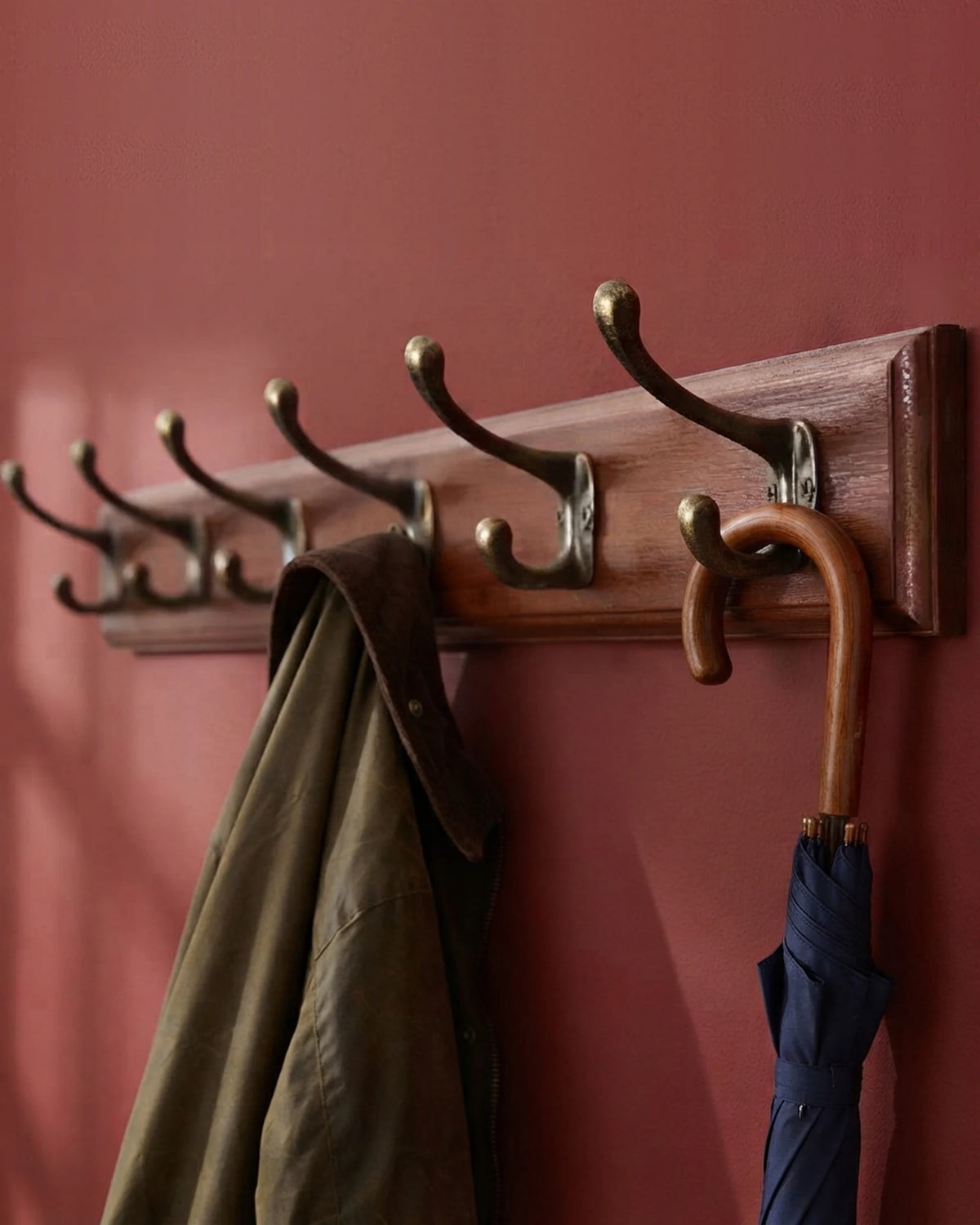 Wooden coat rack with six antique bronze double hooks on a red Victorian hallway wall, holding a waxed jacket and an umbrella